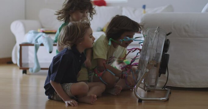 Three children enjoying a cool breeze from an electric fan in a sunny living room