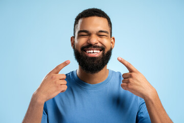 Happy African American man pointing with hands at teeth while closing eyes over blue background