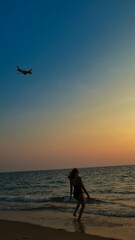 A woman is standing on the beach, watching a plane fly overhead