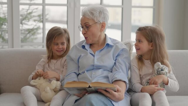 Grandmother with granddaughters resting. Happy relaxed older adult granny embracing reading book with children two twins granddaughters leisure rest on couch at home. Successful, moralizing concept.