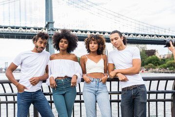 A diverse group of four friends casually dressed, posing confidently with a bridge in the...