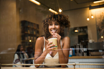 A cheerful young woman with curly hair smiles as she sips on a cold beverage in a cafe, seen...