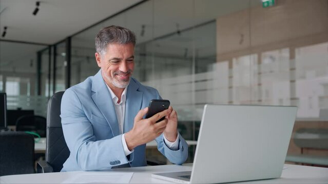 Happy busy middle aged business man ceo investor wearing suit sitting in office using mobile cell phone. Older businessman professional entrepreneur looking at smartphone at work making baking payment