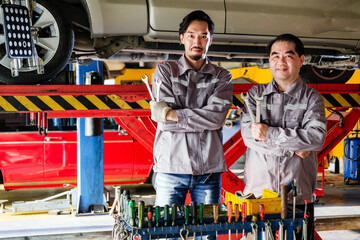 Diversity mechanic teamwork, a Thai and Japanese man in blue uniforms standing and arm crossed in front of the lifted car with working tools. Automobile repairing service. Vehicle maintenance. Labor