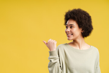 Happy African American woman posing and pointing to copy space with thumb up over yellow background