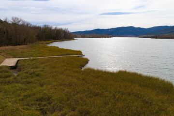 Muelle de madera se adentra en el Lago de Banyoles, con vegetación otoñal y las montañas del Pla de l'Estany como telón de fondo en un día luminoso de invierno. © jocaja