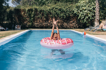 A girl is in a pool with a pink donuts float, she is jumping in the water with arms up and laughing