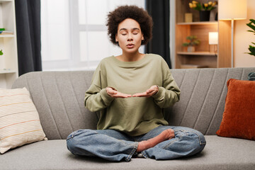 Mindful African American woman breathing out with closed eyes, calming down at home, sitting on sofa