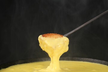 Dipping piece of bread into fondue pot with tasty melted cheese against dark gray background,...