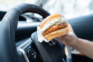 Asian lady holding hamburger to eat in car, dangerous and risk an accident. - Powered by Adobe