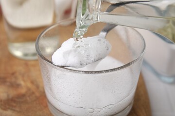 Pouring vinegar into spoon with baking soda over glass bowl at table, closeup