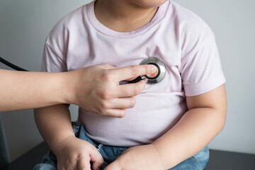 Female doctor checking fat kid put tight shirt lungs during medical checkup at home interior. Friendly female pediatrician using stethoscope to examine breathing and heartbeat of young patient