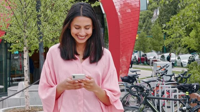 Young smiling happy Indian woman, gen z girl customer standing outdoors on city street or park holding smartphone using mobile cell phone technology dating or shopping apps, chatting in social media.