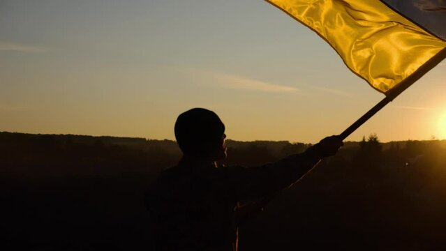 Young man in military uniform waving flag of Ukraine against beautiful sunset at background. Male ukrainian army soldier lifted national banner at countryside. 