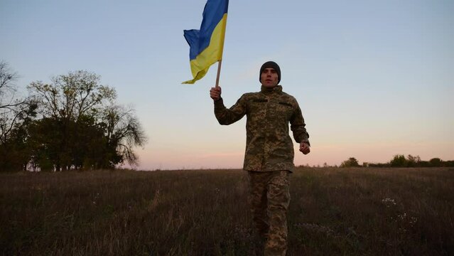 Young male military in uniform jogging with flag of Ukraine on meadow at dusk. Soldier of ukrainian army waving blue-yellow banner in honor of victory against russian aggression. End of war. Slow mo