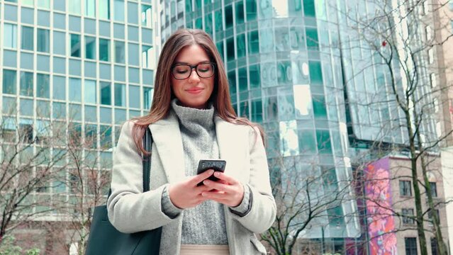Young smiling happy woman, business lady standing outdoors on city street wearing eyeglasses holding smartphone using mobile cell phone technology, texting on cellphone.