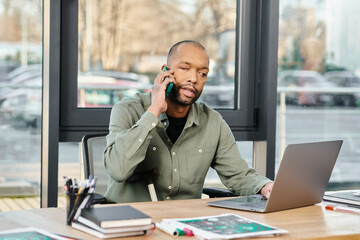 black man sits at a desk, fully engaged on a cell phone call, deep in conversation.