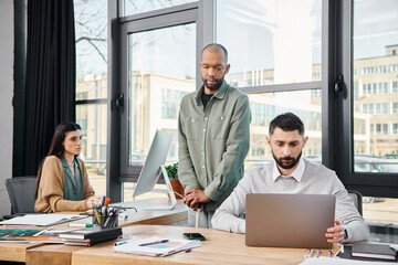 A man focused at a desk, typing on a laptop in a busy office setting, immersed in the corporate culture of a business project.