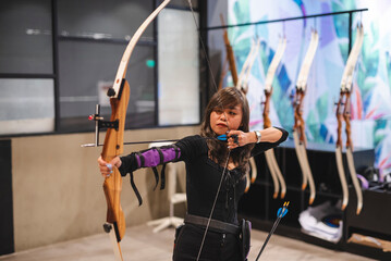 A young Asian woman is captured in a moment of focus as she practices archery at an indoor range located in a shopping mall. - obrazy, fototapety, plakaty