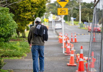 Man walking on a pedestrian footpath. Orange traffic cones and metal fences on the road. Road works in Auckland. - 789140352