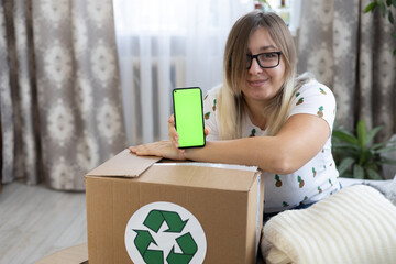 Smiling cute woman holds a phone with a chromakey screen in her hand over a box with a recycling...