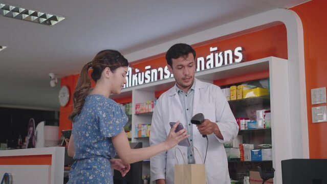 A male pharmacist uses a barcode scanner for smartphone payments in a modern pharmacy, The neon sign in Thai reads "Pharmacist Service Area"