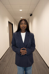 A poised young Black woman stands in an office hallway, her hands clasped in a gesture of...