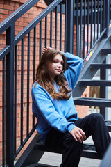 A student girl is sitting in beautiful clothes on an iron staircase. A photo shoot for a girl. - 789175332