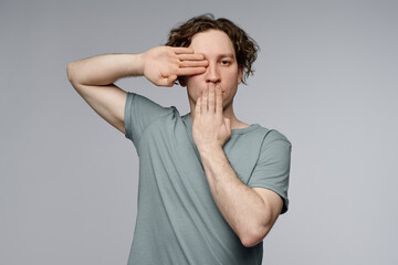 Medium studio shot of young Caucasian man posing for camera covering his eye and mouth with hands,...