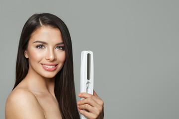 Gorgeous young healthy brunette woman holding hair iron and straightening her long smooth shiny...