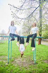 Three girls of high school age in physical education uniforms are sitting on a horizontal bar. One is hanging upside down. - 789178151