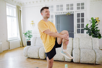 A smiling man stretches before a workout, holding his leg up, in a spacious and bright living room...