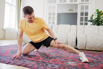 A focused man in sportswear performs stretches on a vibrant rug in a bright, airy room, showcasing...