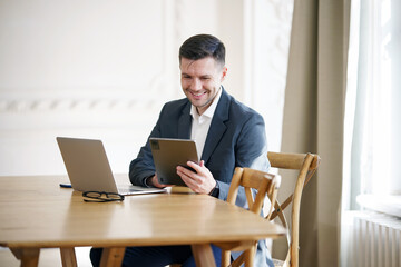 Fototapeta na wymiar An amiable businessman engages with a tablet, grinning as he sits at a wooden table, with a laptop and eyeglasses nearby.