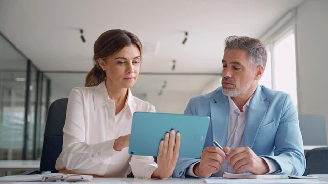 Smiling mature female executive manager holding digital tablet consulting client discussing tech services at corporate meeting. Two happy professional business colleagues working sitting in office.