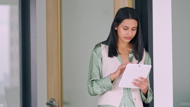 Young smart smiling professional Indian business woman, happy confident female company worker or student holding digital tablet standing in modern office working, looking at tab, copy space.