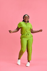Fototapeta na wymiar Young African-American woman in bright green stylish overall dancing of joy against pastel pink studio background. Concept of human emotions, fashion and beauty, trends, hobby, self-expression.