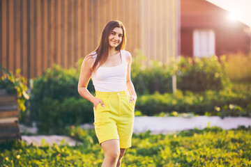 Knee length portrait of woman wearing yellow bermuda shorts, young white female stands against blurred rural background on summer evening before sunset.