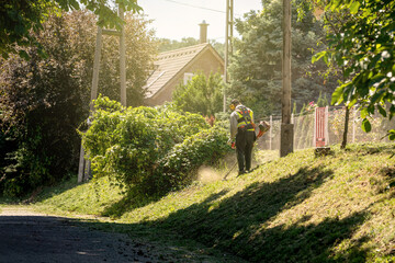 A man mows the grass in the street on the grassy part next to the road. Sunny day. A man in...