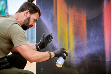 Attractive male artist is painting picture with paint spray can spraying it onto canvas at outdoor...