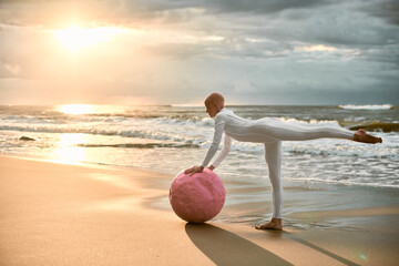 Hairless ballerina with alopecia in white futuristic suit dancing with pink sphere at sunset sea, metaphoric surreal performance exudes confidence, hope and unique beauty