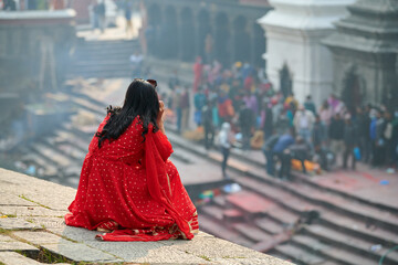 Back view of woman in red sari watching mournful cremation ceremony at Pashupatinath Temple complex, Nepal, view from Pandra Shivalaya site, symbolizing cyclical nature of life and death