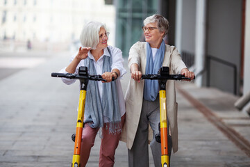 Elderly joyful gray haired women friends ride electric scooters in the city, happy smiling, enjoying a fun time together