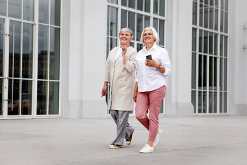 Happy smiling Elderly senior women walk and converse in city, enjoying leisurely stroll together, dressed in stylish fancy clothes, mature female models on street bright background, outdoors