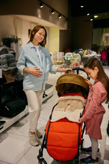 Pregnant mother and little daughter choosing stroller in shopping mall - 789226541