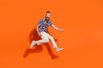 Full length photo of funky eccentric guy wear print shirt white trousers jumping play imaginary guitar isolated on orange color background