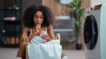 African American female woman girl pensive thinking ponder idea thoughtful think solution hesitate decide using mobile phone online shopping smartphone at floor with laundry basket washing machine - Powered by Adobe