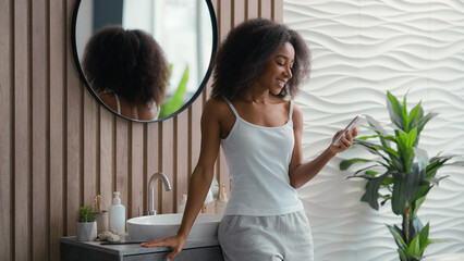African American happy smiling woman using mobile phone browsing chatting smartphone girl telephone social media sms message shopping online internet surfing app in bathroom home beauty procedure