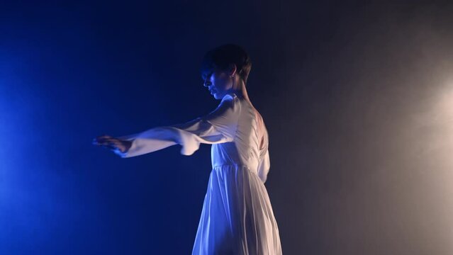 Beautiful elegant woman in white dress performing amazing modern choreography - contemporary style dance with hands in dark studio. Artist performance for advertising dance school, music video.