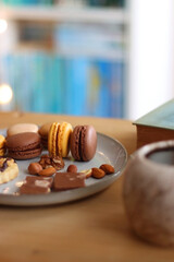 Plate with macarons, cookies, chocolate and nuts, cup of tea or coffee, book and reading glasses on the table. Selective focus. Colorful bookcase in the background.